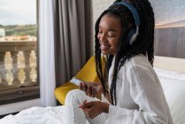 Visão lateral da jovem afro-americana positiva em roupas casuais e fones de ouvido sorrindo enquanto se senta na cama acolhedora com smartphone em casa — Fotografia de Stock