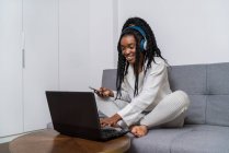 Радісна молода афроамериканська леді з довгим кучерявим волоссям в повсякденному одязі посміхається, слухаючи музику в навушниках з смартфоном і працюючи на комп'ютері, сидячи на дивані вдома. — стокове фото