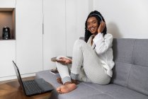 Giovane donna afroamericana gioiosa con lunghi capelli ricci in abiti casual sorridenti mentre ascolta musica in cuffia con smartphone e lavora al computer seduto sul divano di casa — Foto stock