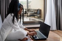 Seitenansicht einer fokussierten jungen ethnischen Freiberuflerin mit langen Afro-Haaren in lässiger Kleidung und Brille, die ferngesteuert am Laptop arbeitet und in einer modernen Wohnung über Kopfhörer Musik hört — Stockfoto
