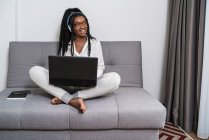 Joven freelancer étnica enfocada con pelo largo Afro en ropa casual y anteojos trabajando remotamente en laptop y escuchando música en auriculares en apartamento moderno - foto de stock