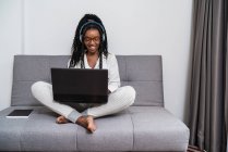 Freelancer jovem e étnico focado com cabelo afro longo em roupas casuais e óculos trabalhando remotamente no laptop e ouvindo música em fones de ouvido no apartamento moderno — Fotografia de Stock