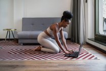 Бічний погляд на щасливу дівчину афроамериканського походження зі спортом посміхається, коли користується ноутбуком, сидячи на підлозі перед онлайн-тренуванням йоги. — стокове фото