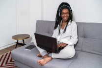 Freelancer jovem e étnico focado com cabelo afro longo em roupas casuais e óculos trabalhando remotamente no laptop e ouvindo música em fones de ouvido no apartamento moderno — Fotografia de Stock