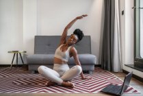 Cuerpo completo de mujer negra joven concentrada en ropa deportiva sentada en la esterilla viendo video en el portátil y realizando pose de yoga durante el entrenamiento de yoga a distancia en casa - foto de stock