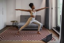 Corpo pieno di concentrato giovane femmina nera in activewear guardare video sul computer portatile e l'esecuzione Virabhadrasana posa durante l'allenamento di yoga a distanza a casa — Foto stock