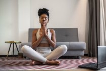 Розслаблена молода афроамериканська жінка в спортивному манері медитуючи в позі Лотоса з закритими очима та руками для молитви під час онлайн-сесії в йозі вдома. — стокове фото