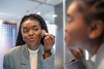 Вид на урожай уверенно молодой афроамериканской девушки с длинными темными волосами, смотрящей в зеркало и наносившей порошок на лицо кистью в ванной комнате — стоковое фото