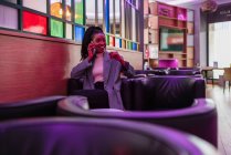Sorrindo jovem afro-americana millennial feminino com longos cabelos encaracolados em roupas da moda sorrindo enquanto descansa em poltrona de couro confortável no café contemporâneo falando no smartphone — Fotografia de Stock