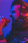 Взрослый бородатый мужчина-певец в капюшоне исполняет выразительную песню с микрофоном во время рок-концерта в неоновом освещении — стоковое фото