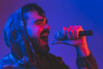 Cantante masculino barbudo adulto con chaqueta encapuchada interpretando una canción expresiva con micrófono durante el concierto de rock en iluminación de neón - foto de stock