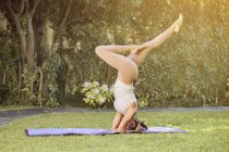 Vista lateral de la feliz hembra flexible en ropa deportiva demostrando la pose de Adho Mukha Vrksasana con las piernas dobladas mientras practica yoga en la esterilla y mirando hacia otro lado - foto de stock