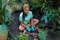 Von oben eine lächelnde schwarze Gärtnerin, die im Gewächshaus auf dem Boden sitzt und Kalanchoe-Blume verpflanzt, während sie in die Kamera blickt — Stockfoto