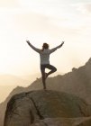 Back view of unrecognizable female standing on top of rocky mountain and doing Tree with Arms Up pose during sunset — Stock Photo