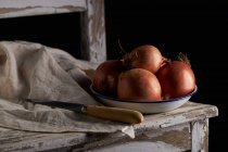Bowl with whole unpeeled onions placed near linen napkin and knife on shabby wooden chair — Stock Photo