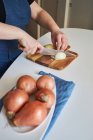 Mulher de colheita em avental cortando cebola crua na tábua de corte na mesa na cozinha em casa — Fotografia de Stock