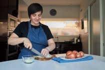 Mujer alegre en delantal sonriendo y picando cebolla cruda en la tabla de cortar en la mesa en la cocina en casa - foto de stock