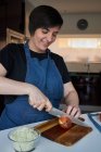 Mulher alegre no avental sorrindo e cortando cebola crua na tábua de corte na mesa na cozinha em casa — Fotografia de Stock