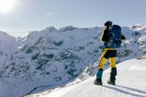 Ganzkörperrückansicht eines nicht wiedererkennbaren männlichen Bergsteigers in warmer Aktivkleidung mit Rucksack, der am Hang des schneebedeckten Felsberges steht und die spektakuläre Landschaft an sonnigen Wintertagen genießt — Stockfoto