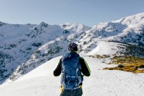 Обратный вид на неузнаваемого альпиниста в теплой спортивной одежде с рюкзаком, стоящим на склоне заснеженной скалистой горы и наслаждающимся впечатляющим пейзажем в солнечный зимний день — стоковое фото