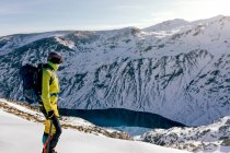 Vue latérale complète du corps de l'alpiniste masculin méconnaissable en vêtements de sport chauds avec sac à dos debout sur la pente de la montagne rocheuse enneigée et profiter d'un paysage spectaculaire dans la journée ensoleillée d'hiver — Photo de stock