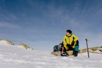 Снизу уверенный в себе альпинист в стильной теплая одежда с рюкзаком альпинистское снаряжение сидит на вершине горы и смотрит в сторону, исследуя снежный скалистый пейзаж в солнечную погоду — стоковое фото