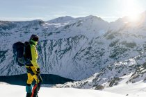 Боковой вид на неузнаваемого альпиниста в теплой одежде с рюкзаком, стоящим на склоне заснеженной скалистой горы и наслаждающимся впечатляющим пейзажем в солнечный зимний день — стоковое фото
