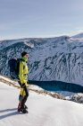 Vista laterale del corpo completo dell'alpinista maschio in caldo activewear con zaino in piedi sul pendio della montagna rocciosa innevata e godendo di un paesaggio spettacolare nella soleggiata giornata invernale — Foto stock