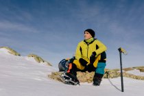Знизу впевнений чоловічий альпініст у стильному теплому активному одязі з рюкзаком обладнання для скелелазіння, що сидить на вершині гори і дивиться далеко, досліджуючи сніговий скелястий пейзаж у сонячну погоду — стокове фото