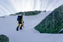 Vista lateral do alpinista irreconhecível andando na encosta da neve coberto cordilheira rochosa em tempo ensolarado — Fotografia de Stock
