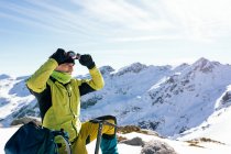 Вид збоку впевненого чоловіка альпініста в стильному теплому активному одязі з рюкзаком обладнання для скелелазіння, що стоїть на вершині гори і дивиться далеко, досліджуючи сніговий скелястий пейзаж в сонячну погоду — стокове фото