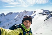 Веселый взрослый альпинист в теплой одежде смотрит в камеру, делая селфи против величественных снежных скалистых вершин в солнечный день — стоковое фото