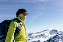 Вид знизу чоловічий альпініст у теплому активному одязі з рюкзаком, що стоїть на схилі засніженої скелястої гори та насолоджується вражаючим пейзажем у сонячний зимовий день — стокове фото