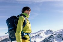 From below side view of male mountaineer in warm activewear with backpack standing on slope of snowy rocky mountain and enjoying spectacular landscape in sunny winter day — Stock Photo