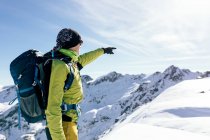 Vista lateral de un montañista masculino irreconocible en ropa deportiva cálida con mochila de pie apuntando a la pendiente de una montaña rocosa nevada y disfrutando de un paisaje espectacular en el soleado día de invierno. - foto de stock
