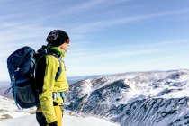 Вид збоку на непізнаваний альпініст у теплому активному одязі з рюкзаком, що стоїть на схилі засніженої скелястої гори та насолоджується вражаючим пейзажем у сонячний зимовий день — стокове фото