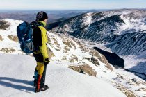 Full body side view of unrecognizable male mountaineer in warm activewear with backpack standing on slope of snowy rocky mountain and enjoying spectacular landscape in sunny winter day — Stock Photo
