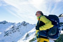 From below side view of male mountaineer in warm activewear with backpack standing on slope of snowy rocky mountain and enjoying spectacular landscape in sunny winter day — Stock Photo