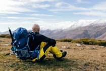 Ganzkörper-Seitenansicht eines erwachsenen aktiven männlichen Bergsteigers in Aktivkleidung mit Rucksack, der sich auf einem grasbewachsenen Berggipfel ausruht und einen malerischen Blick auf die schneebedeckten Berge genießt — Stockfoto