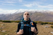 Hombre calvo adulto montañero activo con los ojos cerrados en ropa deportiva con mochila descansando en la cima de la montaña y disfrutando de una pintoresca vista de la cordillera cubierta de nieve - foto de stock