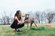 Cheerful female owner embracing loyal purebred Weimaraner dog while sitting together on border on grassy meadow in park — Stock Photo