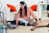 Високий кут повного тіла весела молода жінка дивиться фільм на ноутбук і погладжує собаку Веймаранерів, проводячи вільний час разом у вітальні — стокове фото