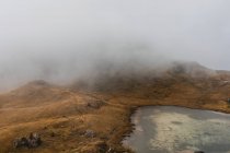 Desde arriba paisaje escénico de pequeño lago rodeado de colinas cubiertas de niebla y nubes en la cordillera de los Dolomitas en Italia - foto de stock