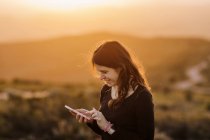 Vue latérale de ravi téléphone portable de navigation féminine tout en se tenant debout sur la colline contre la montagne sous le coucher du soleil — Photo de stock