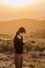 Side view of delighted female browsing mobile phone while standing on hill against mountain under sunset sky — Stock Photo