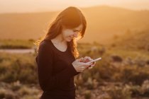 Вид збоку в захваті від жіночого перегляду мобільного телефону, стоячи на пагорбі на горі під небом заходу сонця — стокове фото