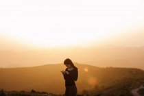 Side view of delighted female browsing mobile phone while standing on hill against mountain under sunset sky — Stock Photo