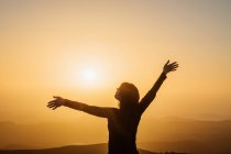 Вид ззаду на невпізнавану жінку, що стоїть з піднятими руками на пагорбі і насолоджується свободою, милуючись гірськими пейзажами на заході сонця — стокове фото