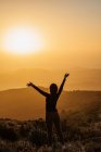 Back view of unrecognizable female standing with raised arms on hill and enjoying freedom while admiring mountainous scenery at sunset — Stock Photo