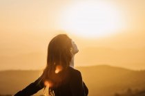 Side view of happy female standing with eyes closed on hill and enjoying freedom while admiring mountainous scenery at sunset — Stock Photo